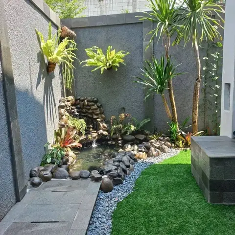 Kolam kecil dengan air terjun mini, dikelilingi batu hitam dan kerikil putih di samping dinding abu-abu