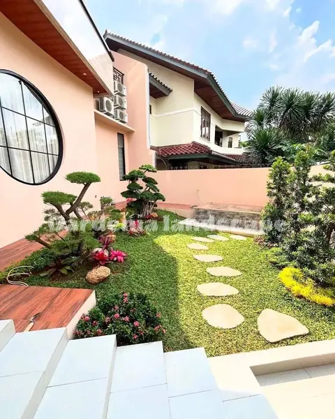 Taman rumput dengan batu pijakan pipih dan bonsai kecil di samping rumah berwarna merah muda