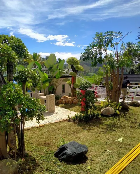 Taman berumput dengan batu hitam besar, jalan batu, dan aneka pohon di bawah langit biru
