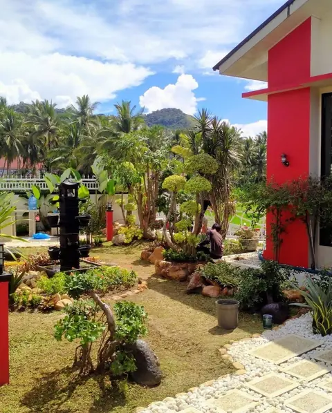 Taman luas dengan batu, bonsai, dan jalan batu putih di samping rumah berdinding merah
